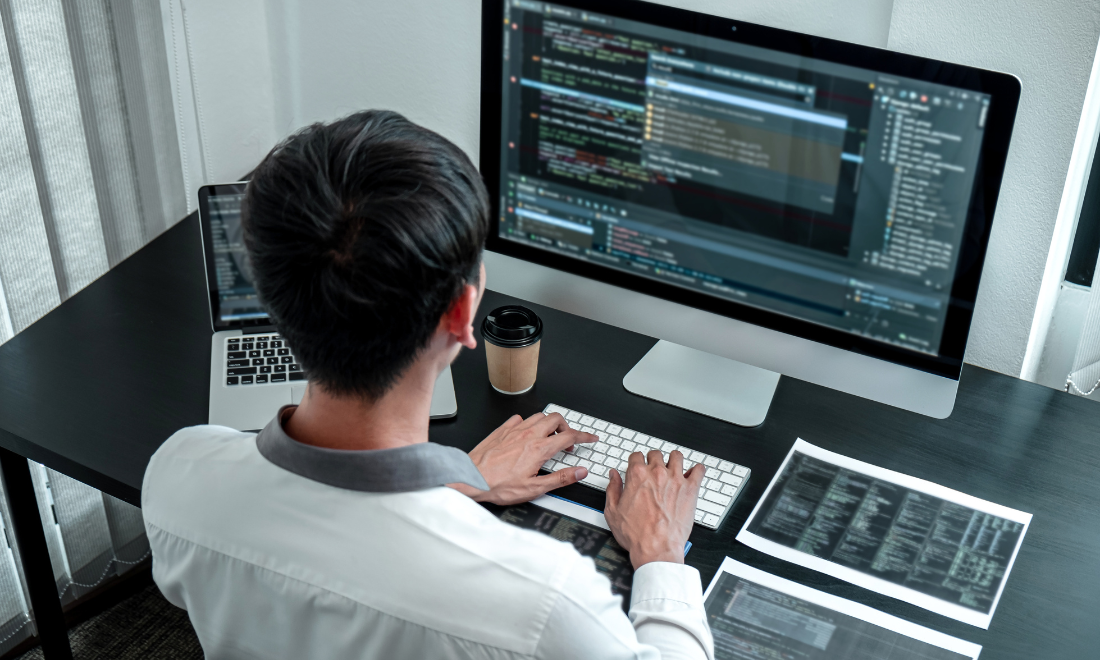 An MLOps engineer works at a desk with a large monitor, laptop, and printed code sheets in a modern workspace.