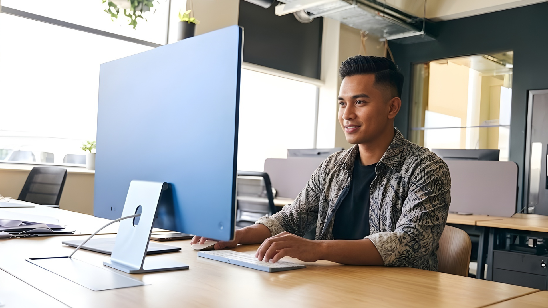 A remote salesforce developer malaysian smiling while working on a desktop in an open-plan office