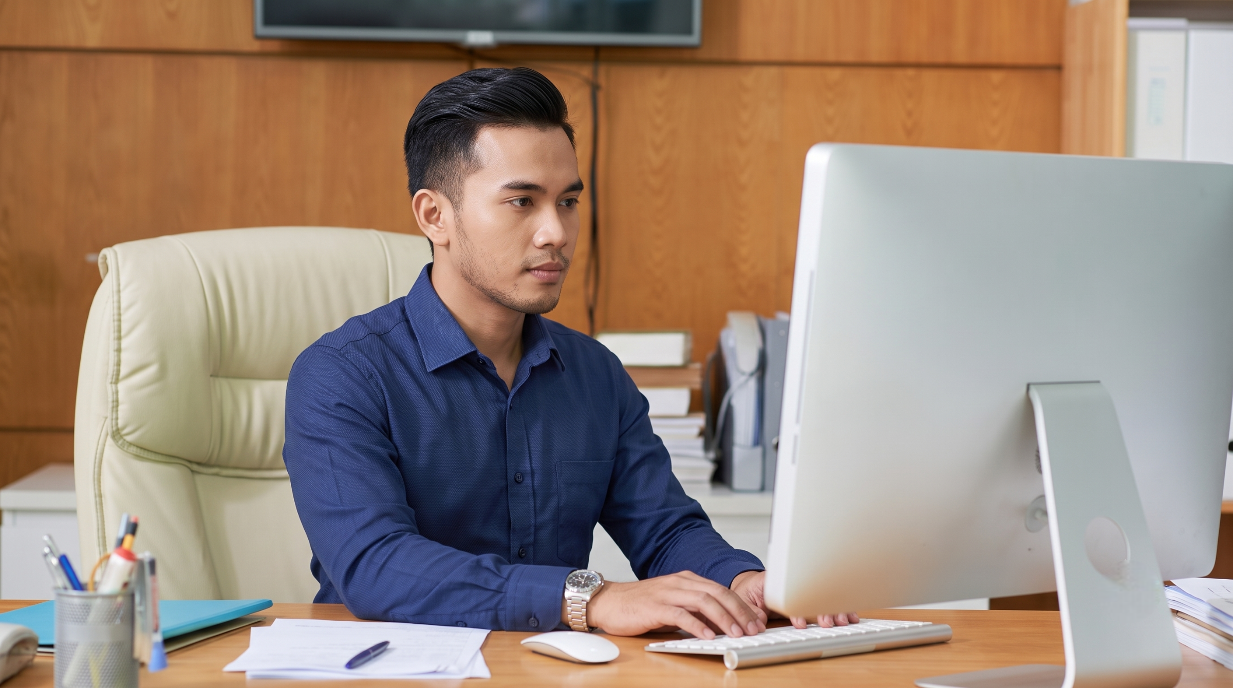 malaysian remote payroll officer typing on a desktop computer at his desk