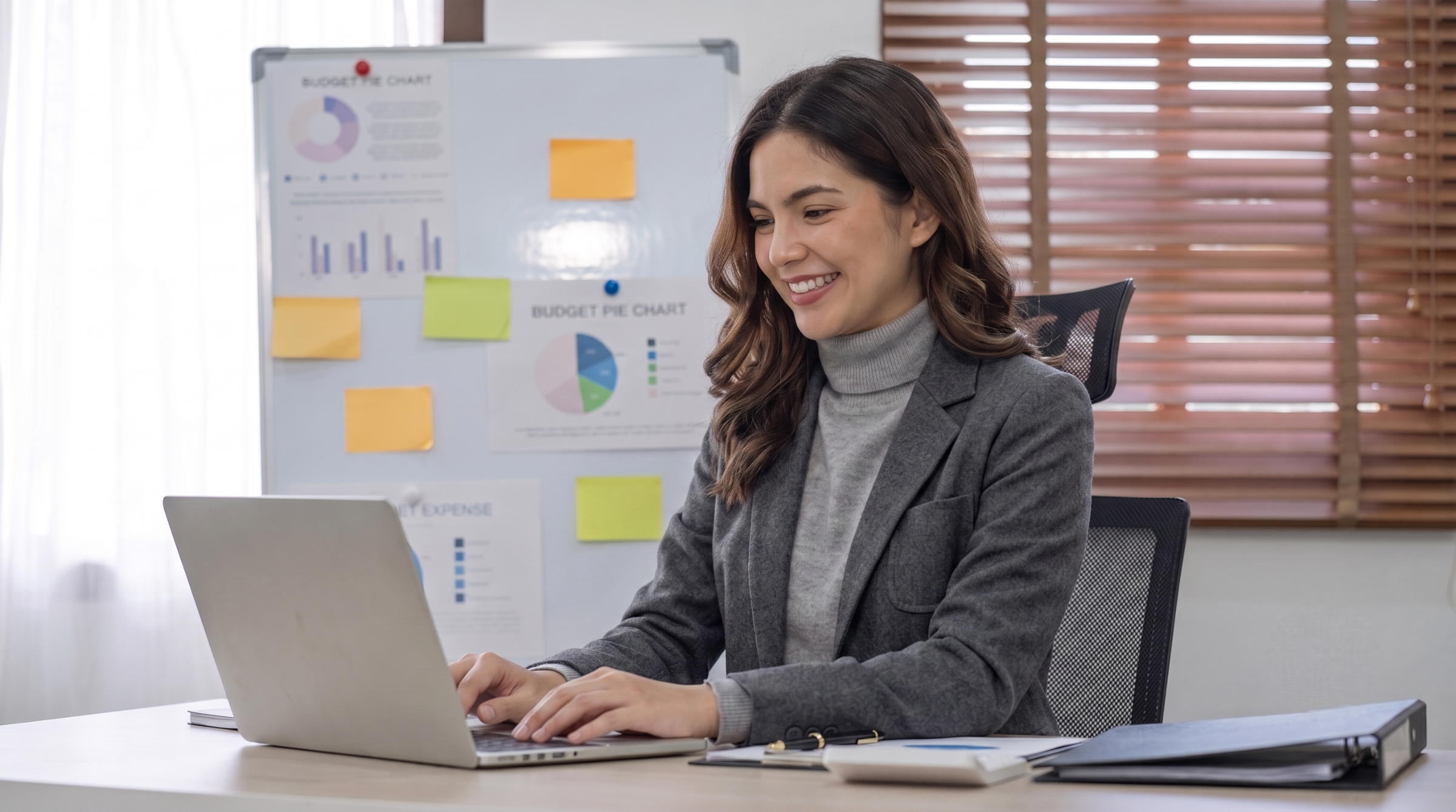 outsourced marketing assistant smiling while typing on her laptop at a desk with financial charts on a whiteboard