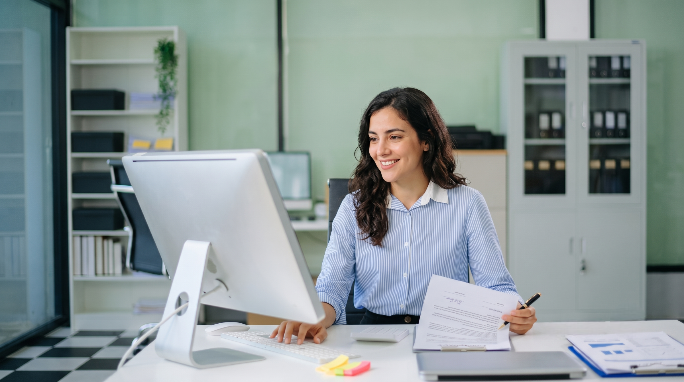 Smiling female remote contract administrator reviewing business paperwork while working on a computer setup