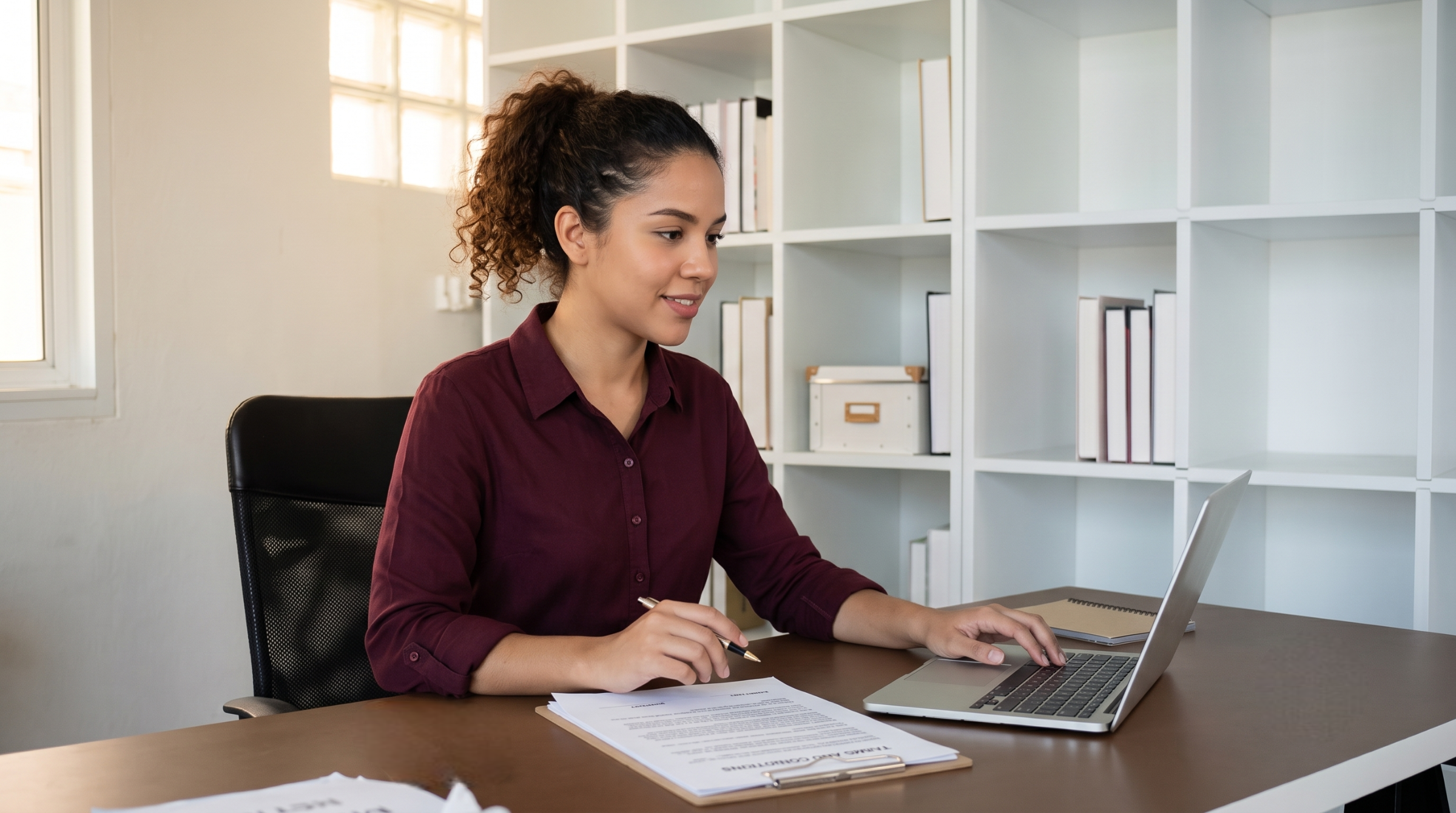 Dedicated remote contract administrator typing on a laptop with agreement documents placed on her desk