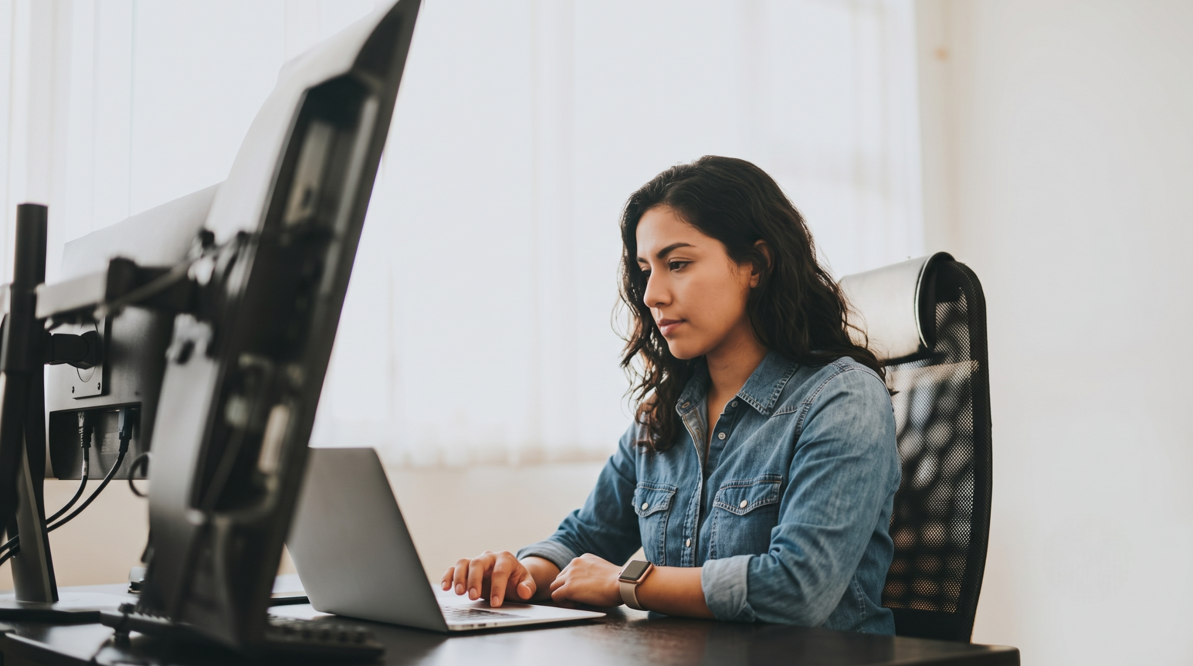latina professional focused on a laptop screen next to a monitor setup, serving as a remote integration engineer in Colombia