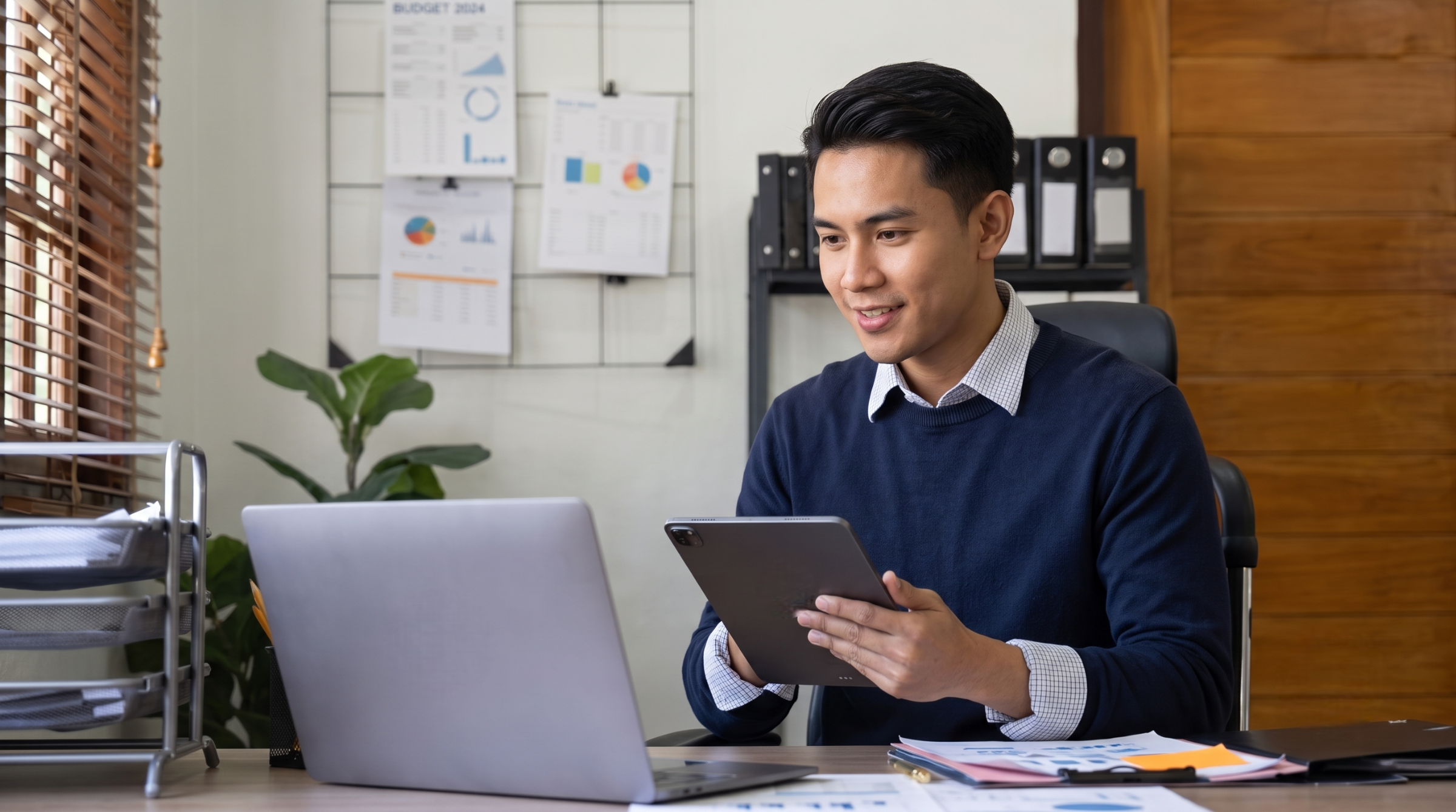 remote marketing assistant in smart casual attire holding a tablet while looking at a laptop screen in an office