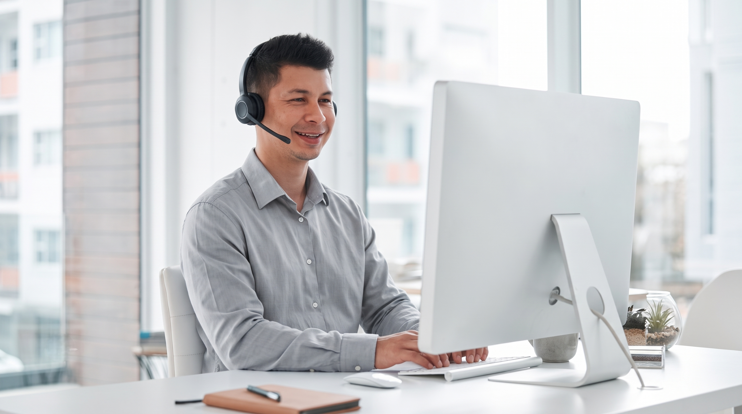Malaysian male remote service coordinator wearing a headset and working at a computer in a modern office setup