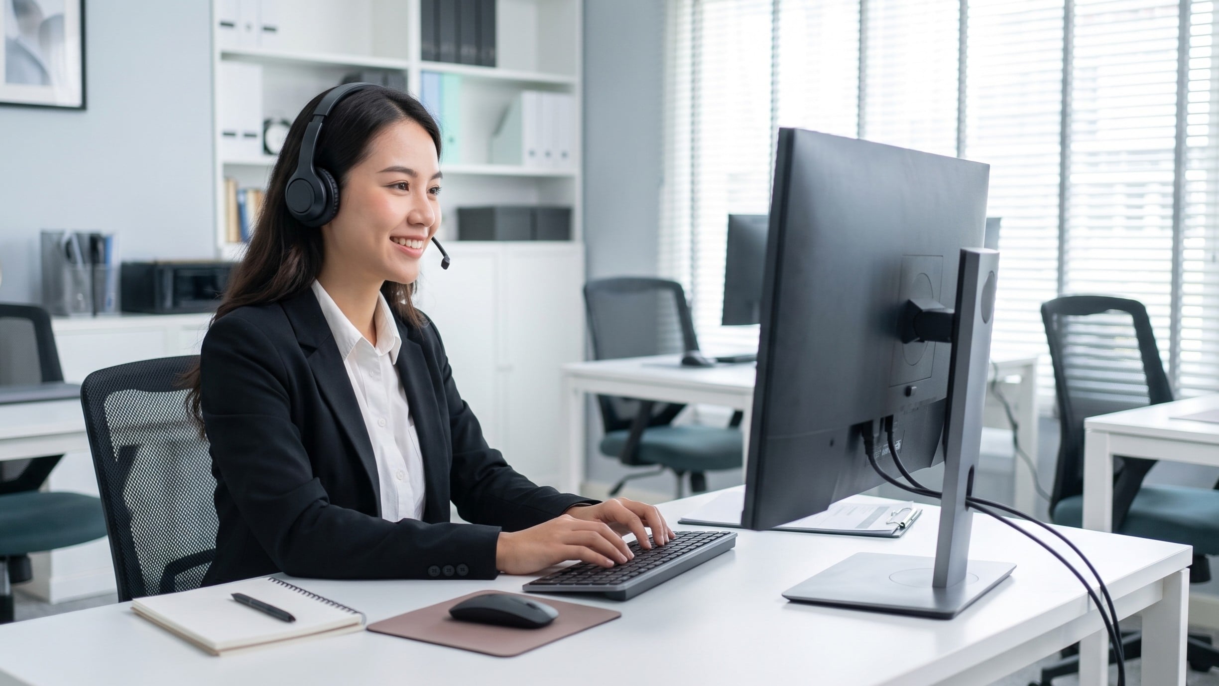 vietnamese woman in a professional suit, using a headset and typing on a computer, demonstrating the duties of a remote member services specialist