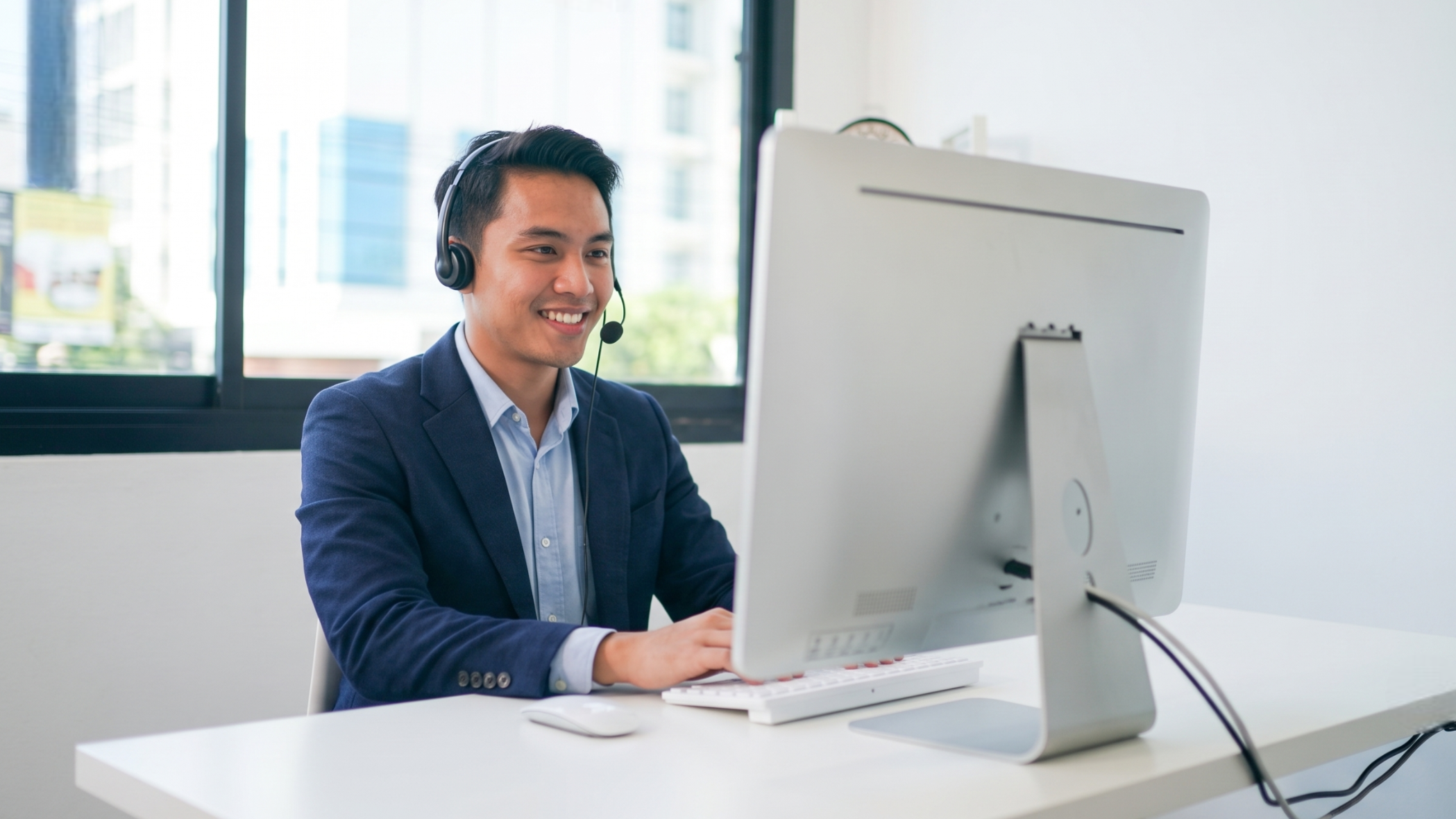 male remote customer experience officer wearing a navy blazer and headset, happily typing on a laptop to assist clients from his home office