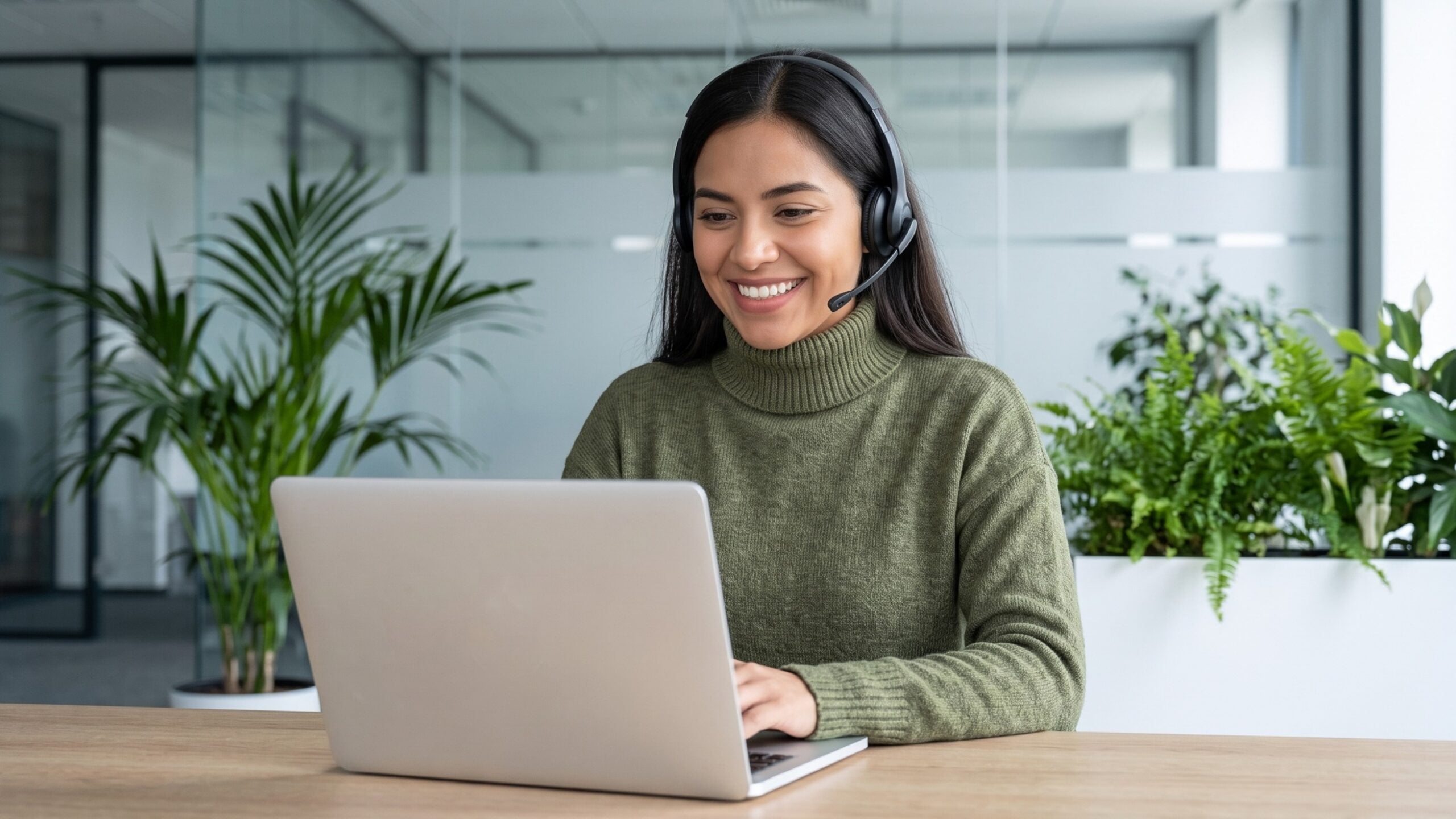 smiling dispatch coordinator remote with a headset using a laptop at a desk surrounded by plants