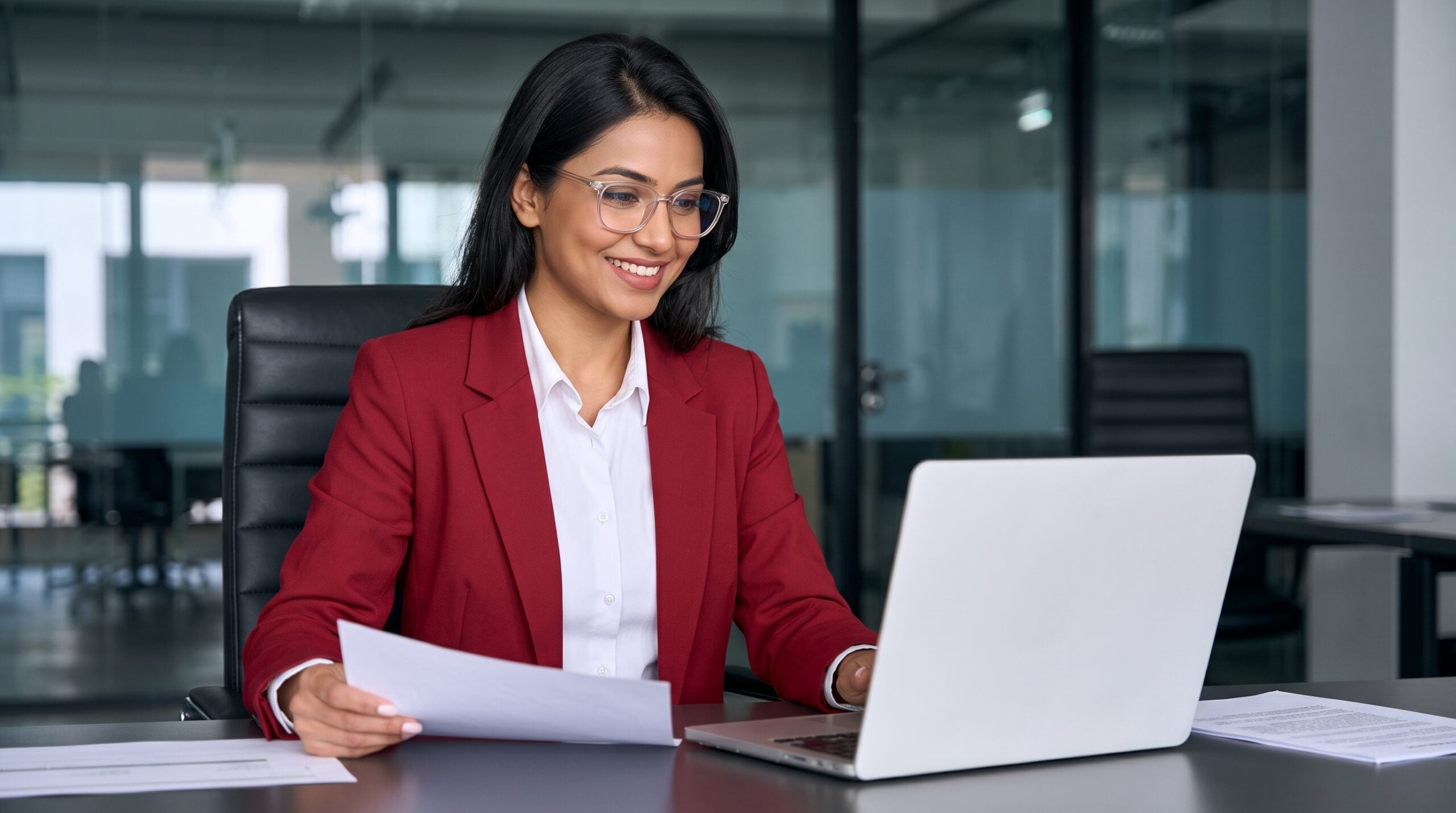 smiling woman in a red blazer and glasses holds a paper and types on her laptop, working from a modern glass office as a remote assistant accountant