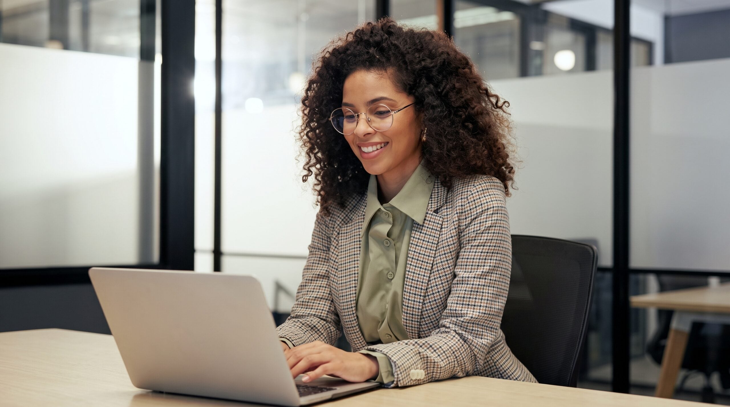 outsourced document controller with curly hair and glasses working on a laptop at a desk in a modern office