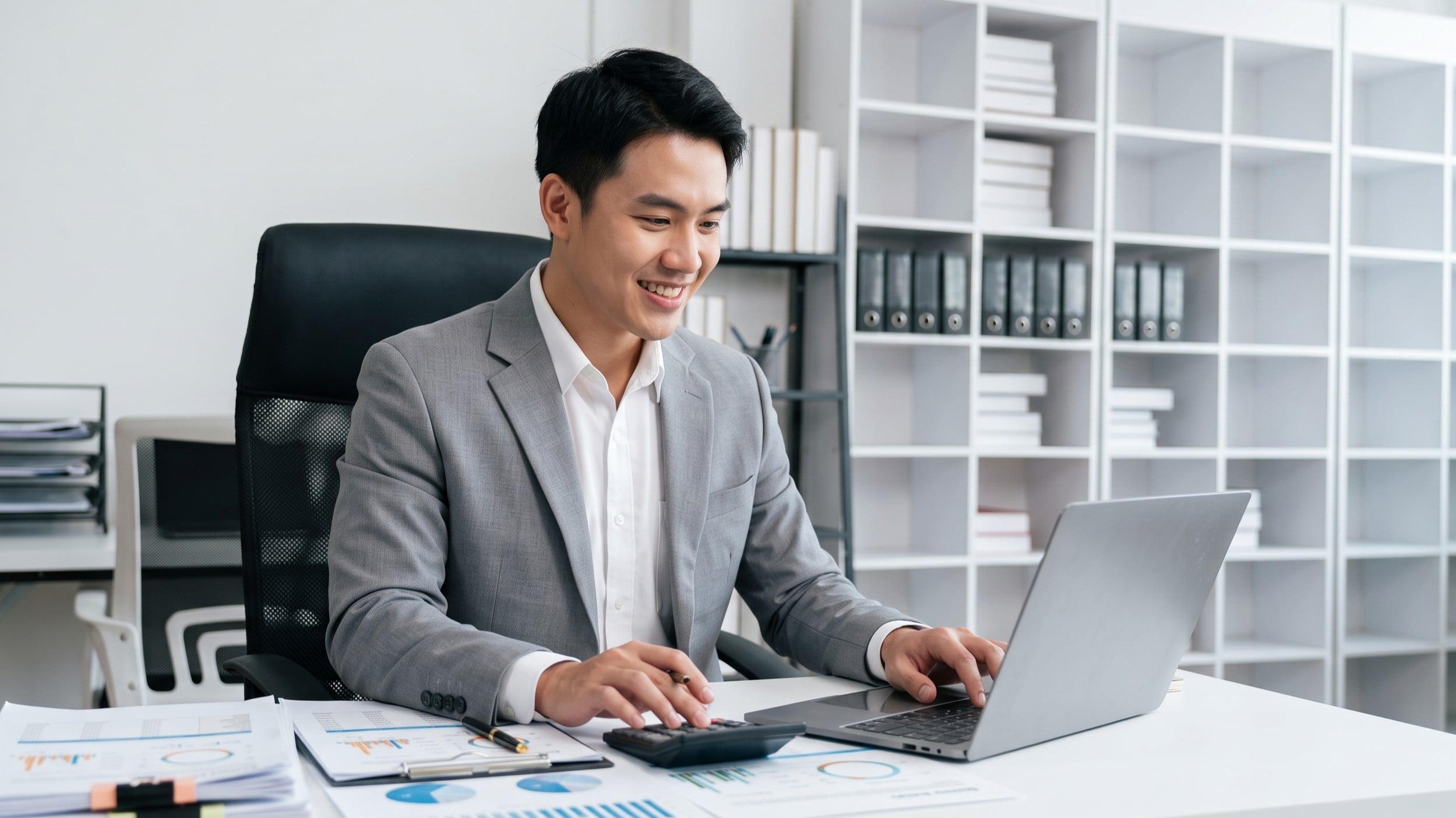 vietnamese male uses a calculator and laptop, working as a virtual financial auditor with files in the background