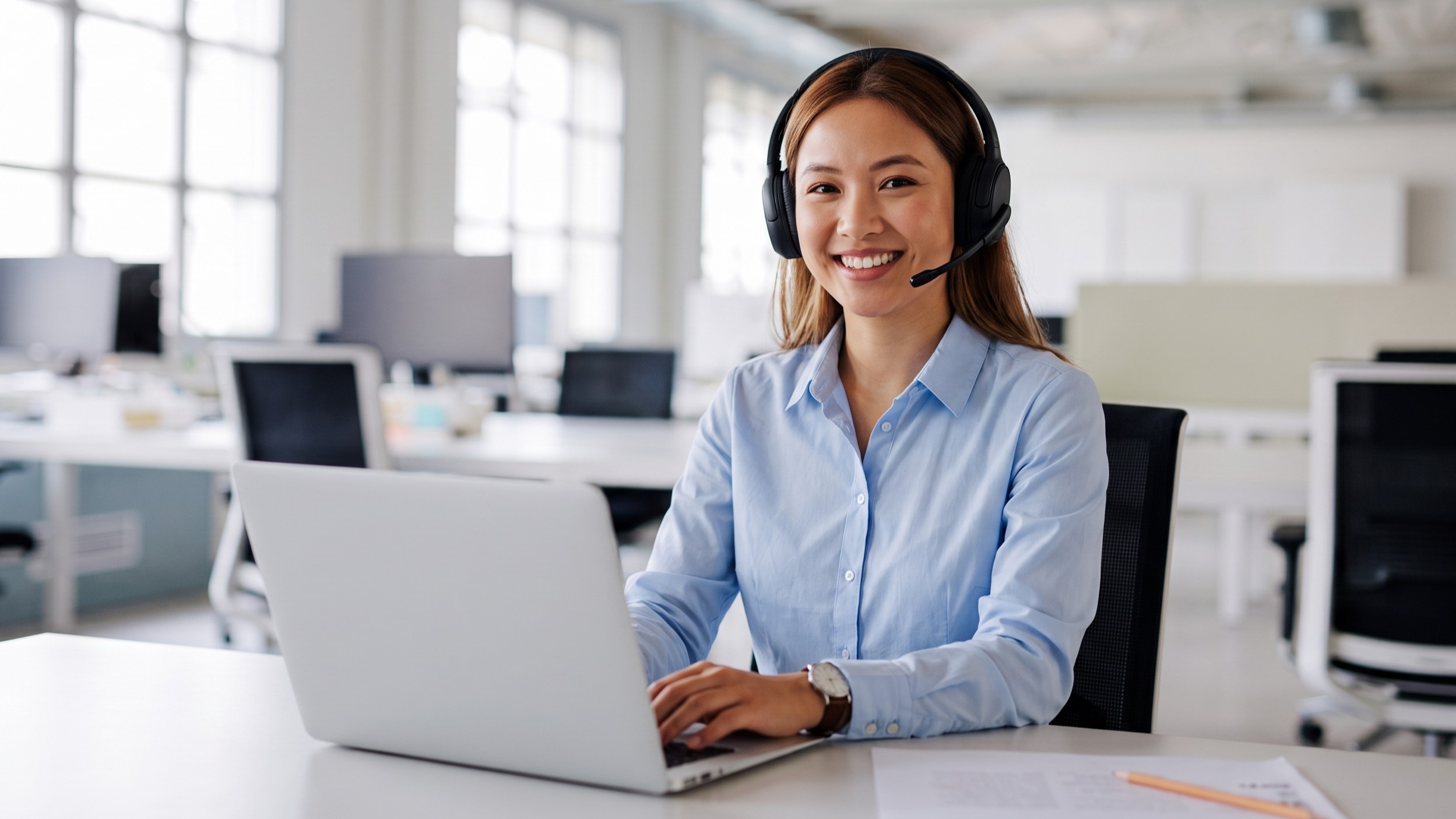 remote Telecommunications Specialist in a light blue shirt, smiling, and typing on a laptop in an office environment.