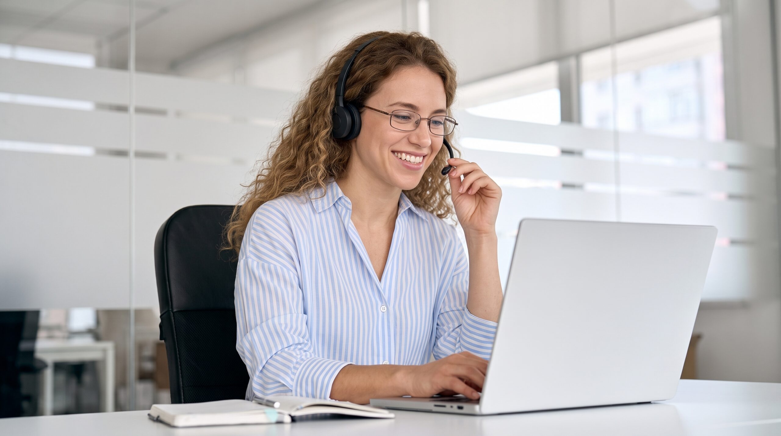 remote Telecommunications Specialist in a striped shirt, smiling and using her laptop while wearing a headset