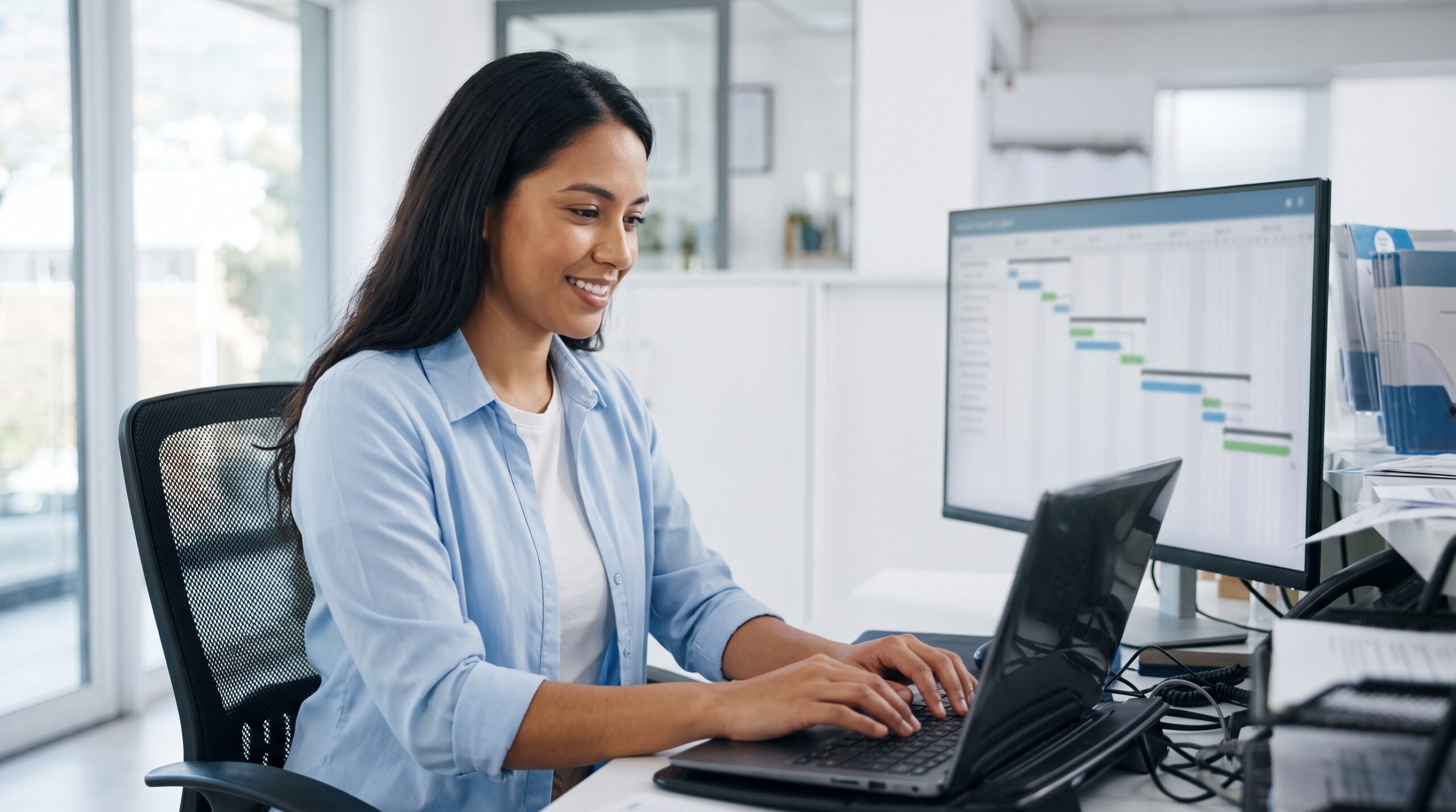 remote implementation manager typing on a laptop with a smile at a desk, with a progress chart on a second monitor in the background